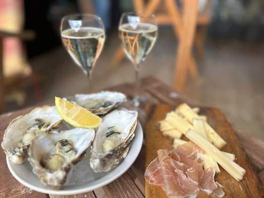 Zwei Gläser Crémant mit Austern, Schinken und Käse auf einem Holzbrett